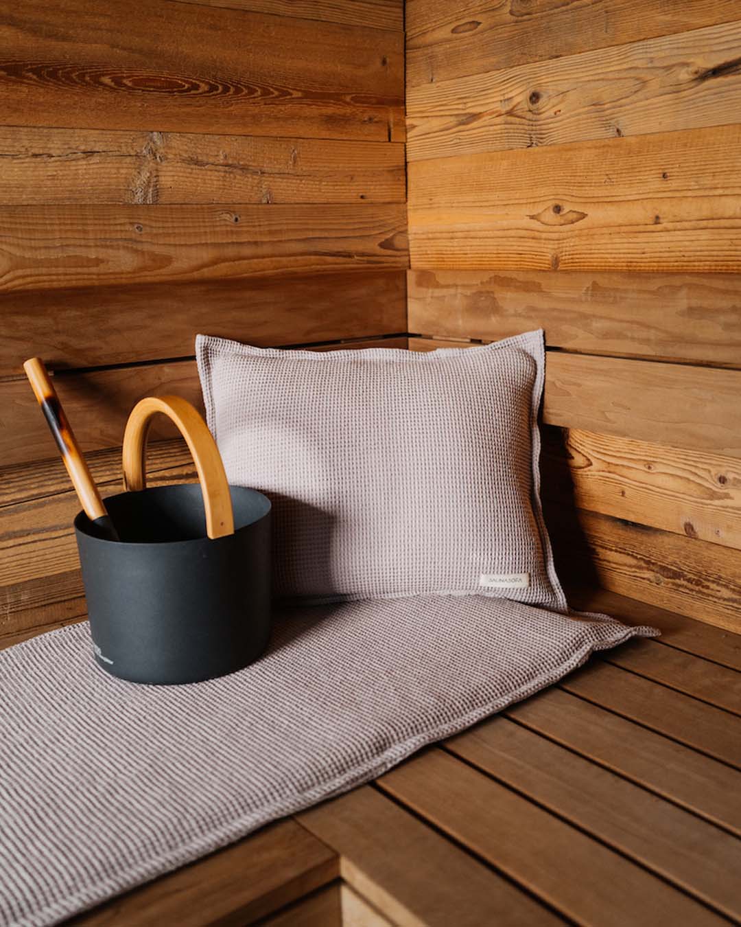 Nahaufnahme von einem Saunakopfkissen und einer Saunaauflage in der Farbe Taupe . Daneben steht der schwarze Badesofa Saunakübel mit Kelle für Aufgüsse. $ Farbe Taupe