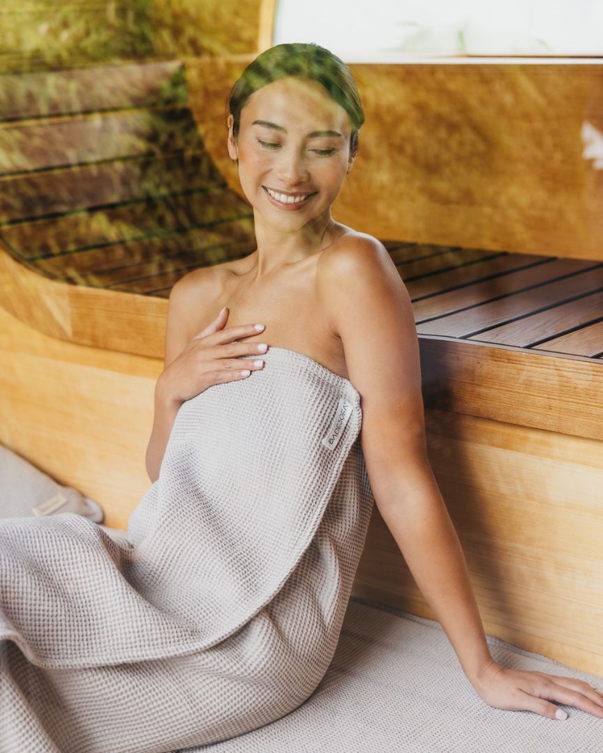 Frau sitz in ein Saunatuch aus Wafflepiqué gewickelt in der Sauna und lächelt.