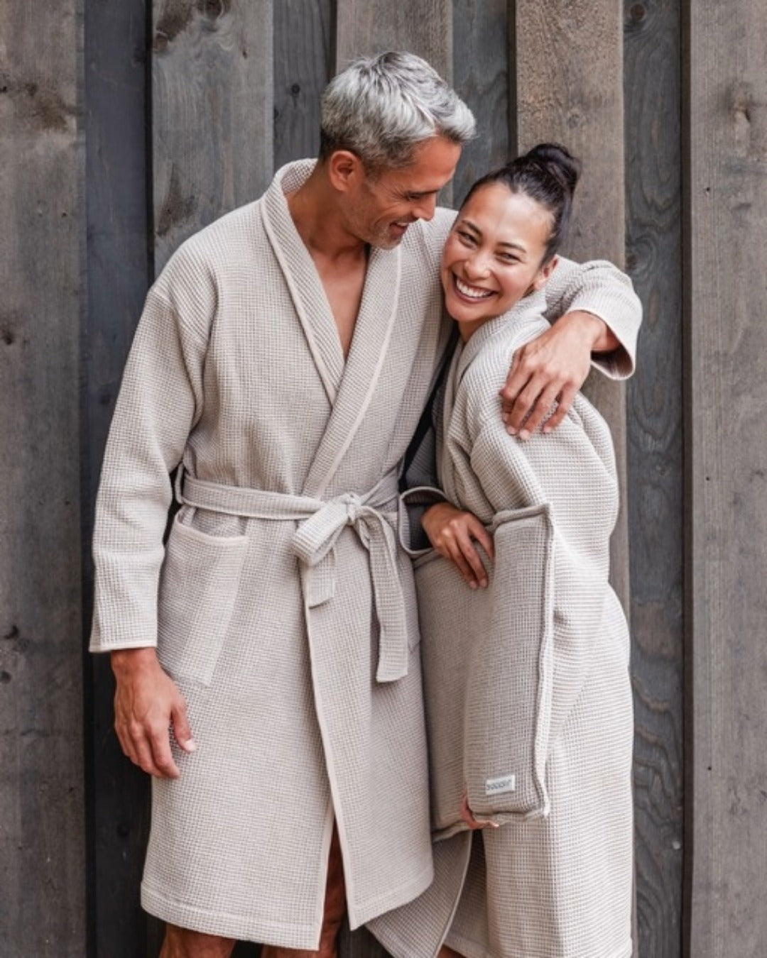 Mann und Frau stehen im Bademantel (Farbe Anthrazit) vor einer Wand und lachen entspannt. | $ai_de::KI generiert | $ai_en::AI-Assisted Creation