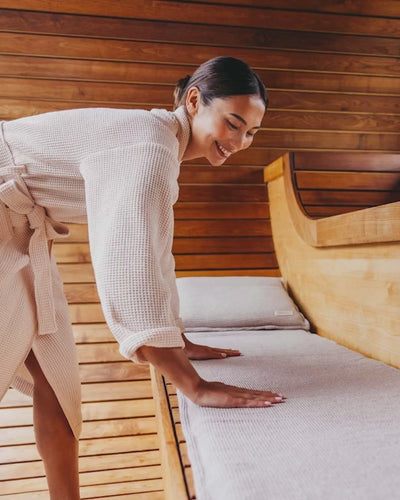 Das Video zeigt, wie eine Frau ihr Saunasofa Set in der Sauna ausrollt, die MAtte glatt streiche und sich dann entspannt hinlegt und den Kopf auf das Kopfkissen legt.